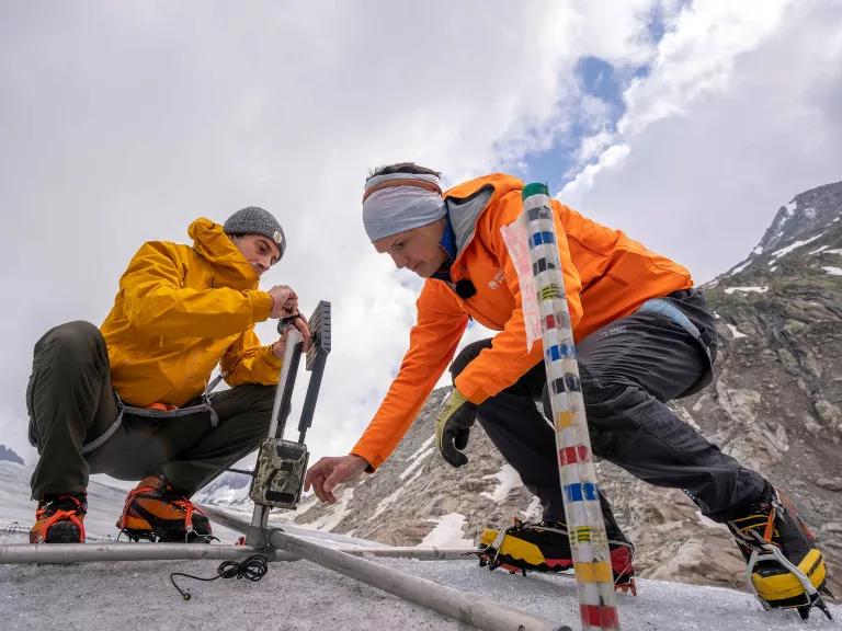 Two people in snow gear crouching over a camera contraption with snowy mountains in the background