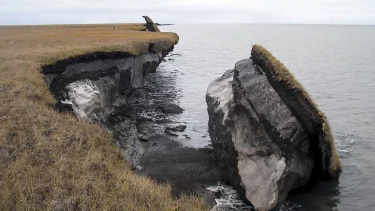 A large chunk of earth broken off of the shore site in water below it, with layers of grass, rock, and ice visible