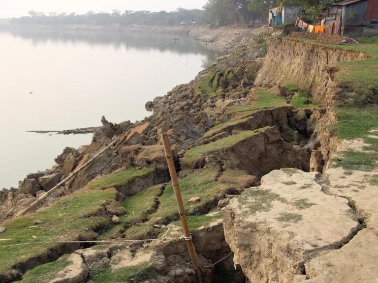 A house sits near the edge of a partially-collapsed river bank, with low grass and dry soil on the surface