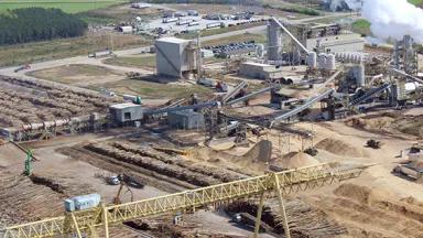 An industrial processing site with thousands of logs stacked in rows outside of a facility with smoke rising from large stacks