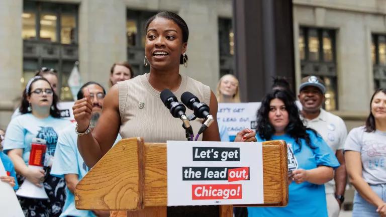 A woman speaking at a podium that has a sign that reads “Let’s get the lead out, Chicago!” with people standing behind her