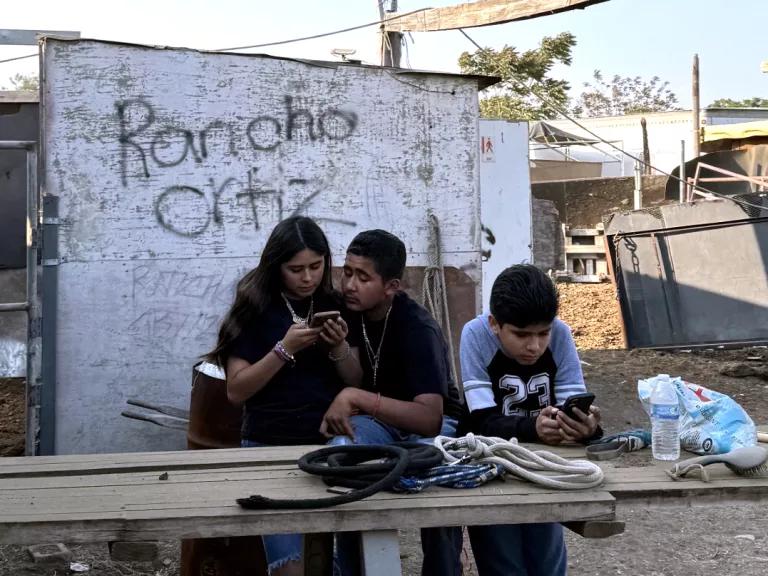 Children leaning against a wooden table outdoors, behind them a rickety wooden wall with the words “Rancho Ortiz” written on it; a child in a pink shirt with their back to the camera in the foreground.