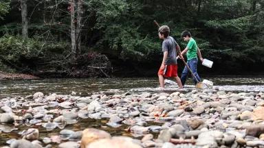 A low angle view of two young boys carrying a net into a river with low water levels, rocks in the foreground