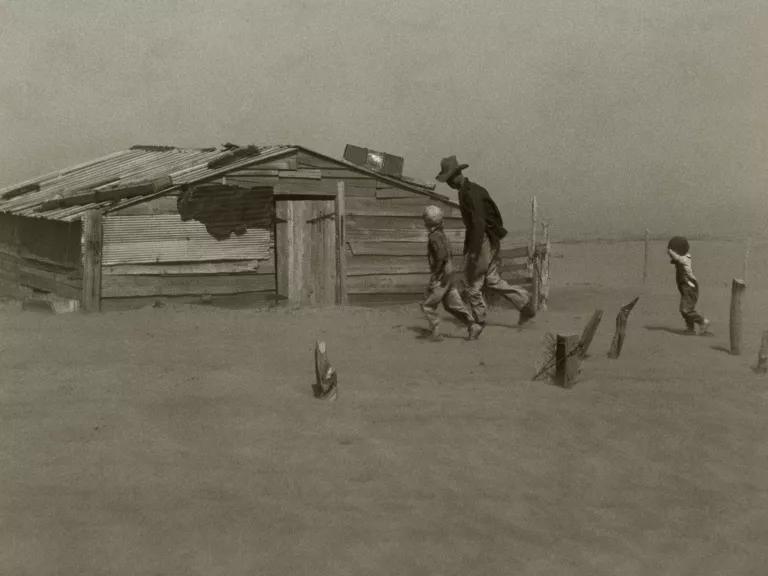 A black and white image shows a man and two children walking in a barren landscape toward a house that appears to be surrounded be buried 2 to 3 feet in dry dirt