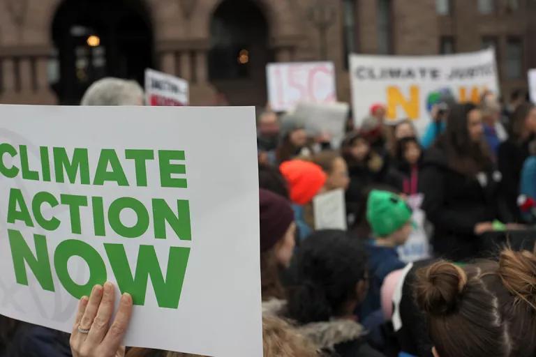 A hand holding up a sign that reads "Climate Action Now," with a crowd in the background