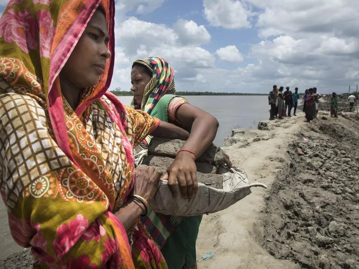 Two women carry containers full of dirt near an embankment