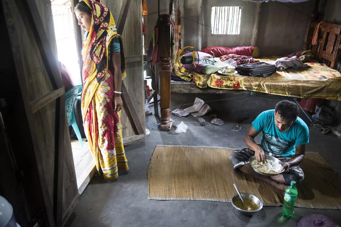 A man sits on a mat on the floor of a room while a woman stands at the doorway looking outside