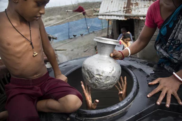 A person's hands emerge from the top of a large black tank as a woman hands them a jug