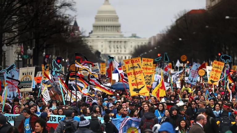 Indigenous water protectors hold up signs and banners during a large protest against the Dakota Access Pipeline (DAPL). The U.S. Capitol building is visible in the distance.