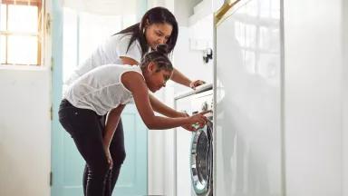 A Black mother and teenage daughter leaning over to press buttons on a washing machine in their home.