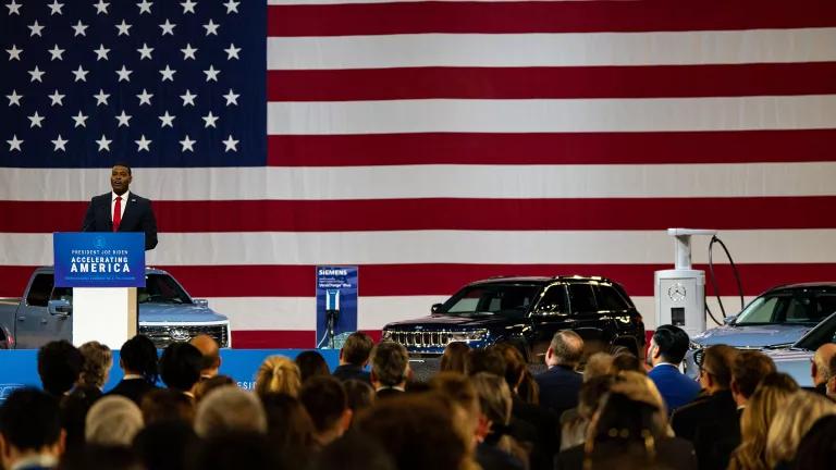 Michael Regan speaking in front of a crowd from a podium on a stage surrounded by electric vehicles (a truck, an SUV, and a sedan) and electric vehicle charging stations, set against a large U.S. flag.