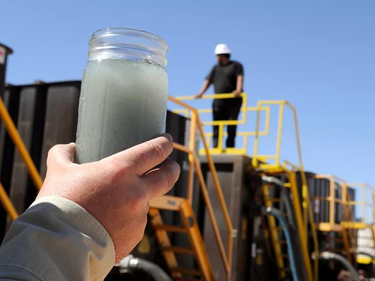 A person holds a glass jar full of dark, murky liquid, with industrial equipment in the background