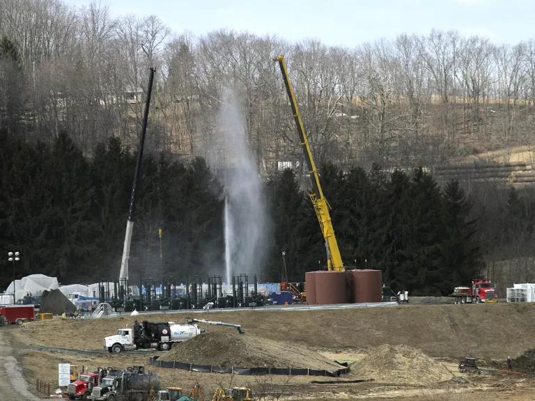 A substance sprays upward from a large gas well, with trees and low mountains in the background