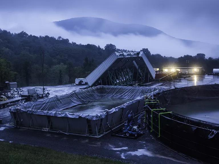 A large rectangular basin lined with sheeting and holding liquid sits in a clearing in front of a mountain in the background