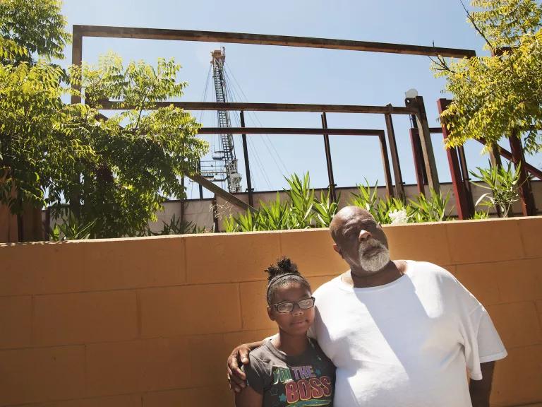 A man and girl stand in front of the cinderblock wall of a garden with plants and small trees, and an oil rig standing tall in the background