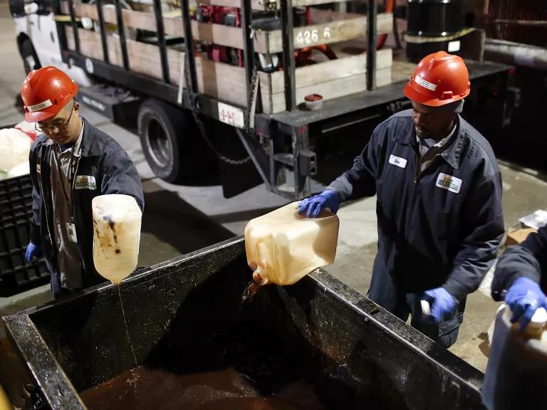 Workers in hard hats pour liquids from plastic containers into a large receptacle