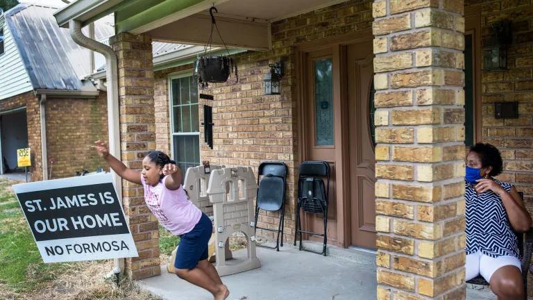 A child plays on the front porch of a brick home while a woman wearing a face mask looks on. A sign on the front lawn reads "St. James is our home. No Formosa"