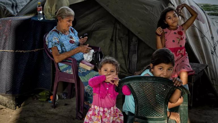 An older woman sits outside of a tent with children playing around her