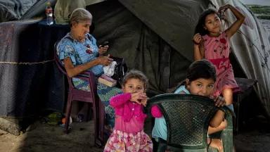 An older woman sits outside of a tent with children playing around her