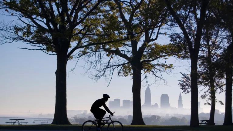 A cyclist rides past trees on a hazy day with a city skyline in the distance