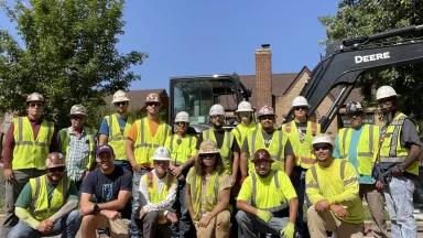 A group of people in hard hats and fluorescent construction vests posing outside of a house