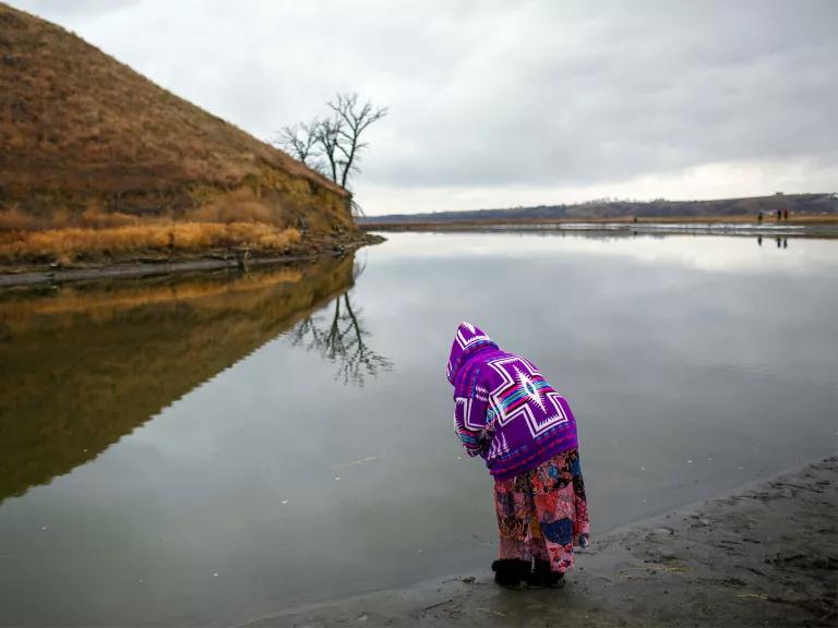 A person stands on the bank of a calm river under a cloudy sky.