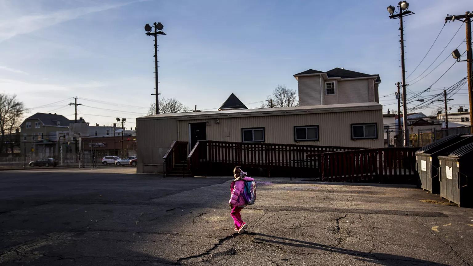 A young girl in a bright pink jacket walks in a paved city parking lot