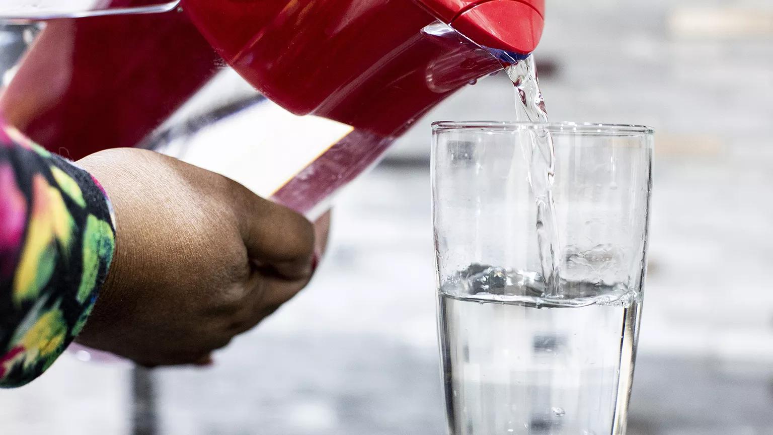 A close-up of a hand pouring water into a glass out of a red pitcher