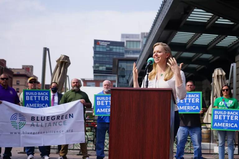 A woman stands at a lectern outside on a city street with a group of people behind her holding signs that read "Build Back Better for America"