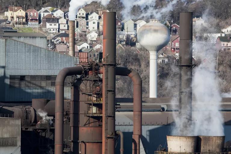 Gray and black smoke rise out of stacks on an industrial facility, with homes visible in the background.