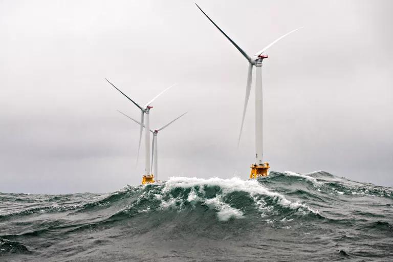 Three wind turbines stand in rough seas with a wave cresting in front of them.