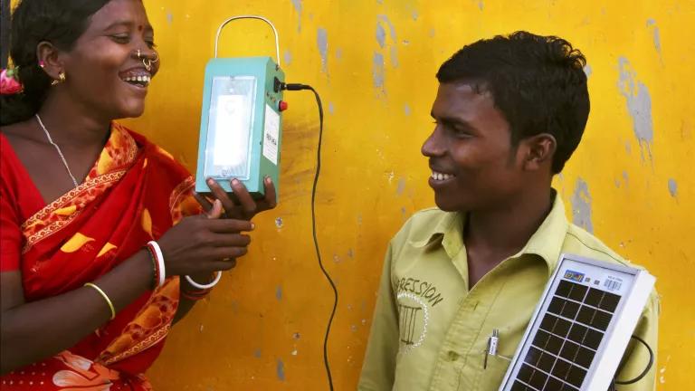 A woman holds a lantern that is connected by a wire to a small solar panel held by a man to her left.