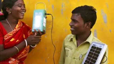 A woman holds a lantern that is connected by a wire to a small solar panel held by a man to her left.