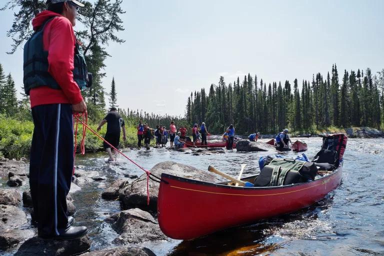 A young man stands on a rocky shore holding a rope attached to a red canoe on the water in front of him