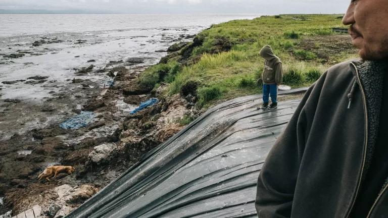 A man and a young boy stand on a grassy ledge partially covered by a large tarp that extends down into the water below