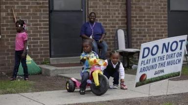 A mother sits outside of her apartment door as her two young sons play, one on a tricycle and the other with a toy car. The mother’s young daughter walks away past a rake leaning against the building. A sign in the dirt reads, “Do not play in the dirt or around the mulch. —EPA.”