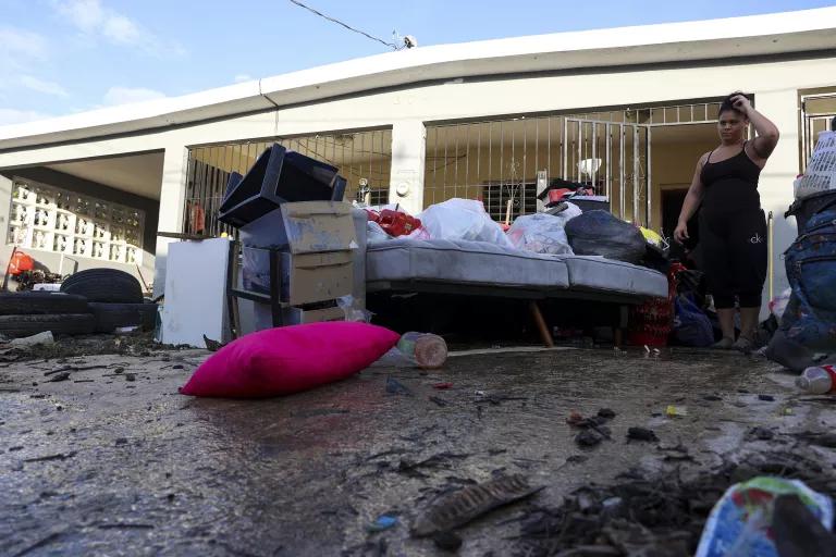 A woman stands near a pile of debris on a muddy sidewalk in front of a house