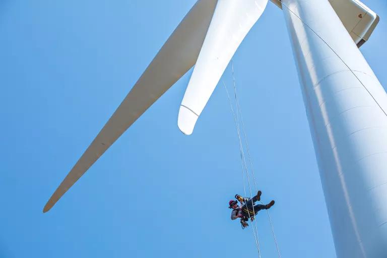 A worker in climbing gear hangs below the blades on a wind turbine