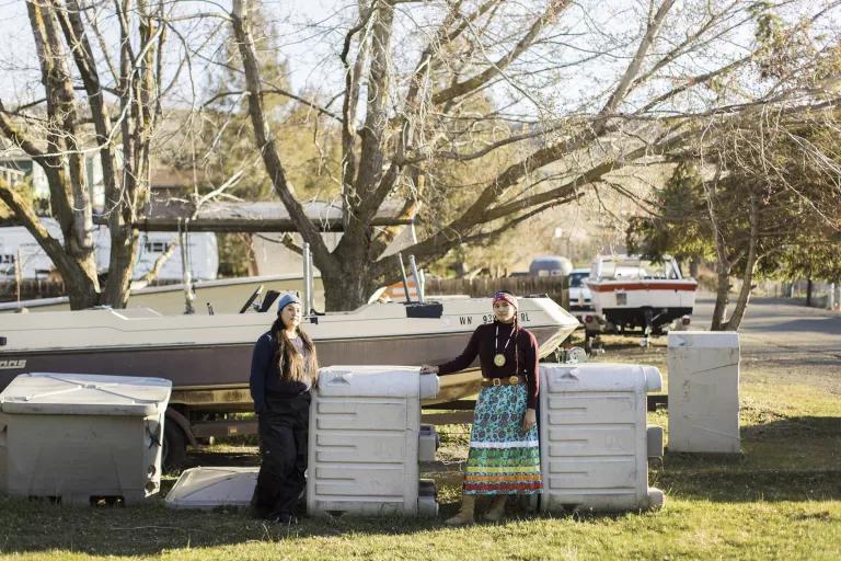 Two women stand next to large equipment and a small boat grounded on the grass behind them