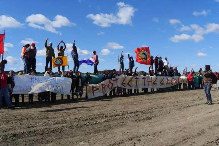 Protesters stand on top of a long stretch of a large above-ground pipe and hold banners and posters