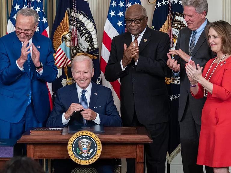 President Joe Biden sits at a small wooden desk and is surrounded lawmakers