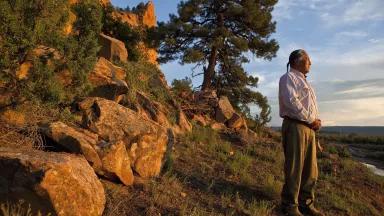 A man stands on a mountain ledge overlooking a vast green valley