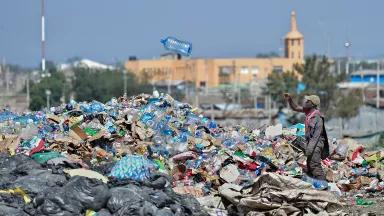 A Black man throwing a large empty bottle of wall onto a pile of plastic and trash that is taller than him